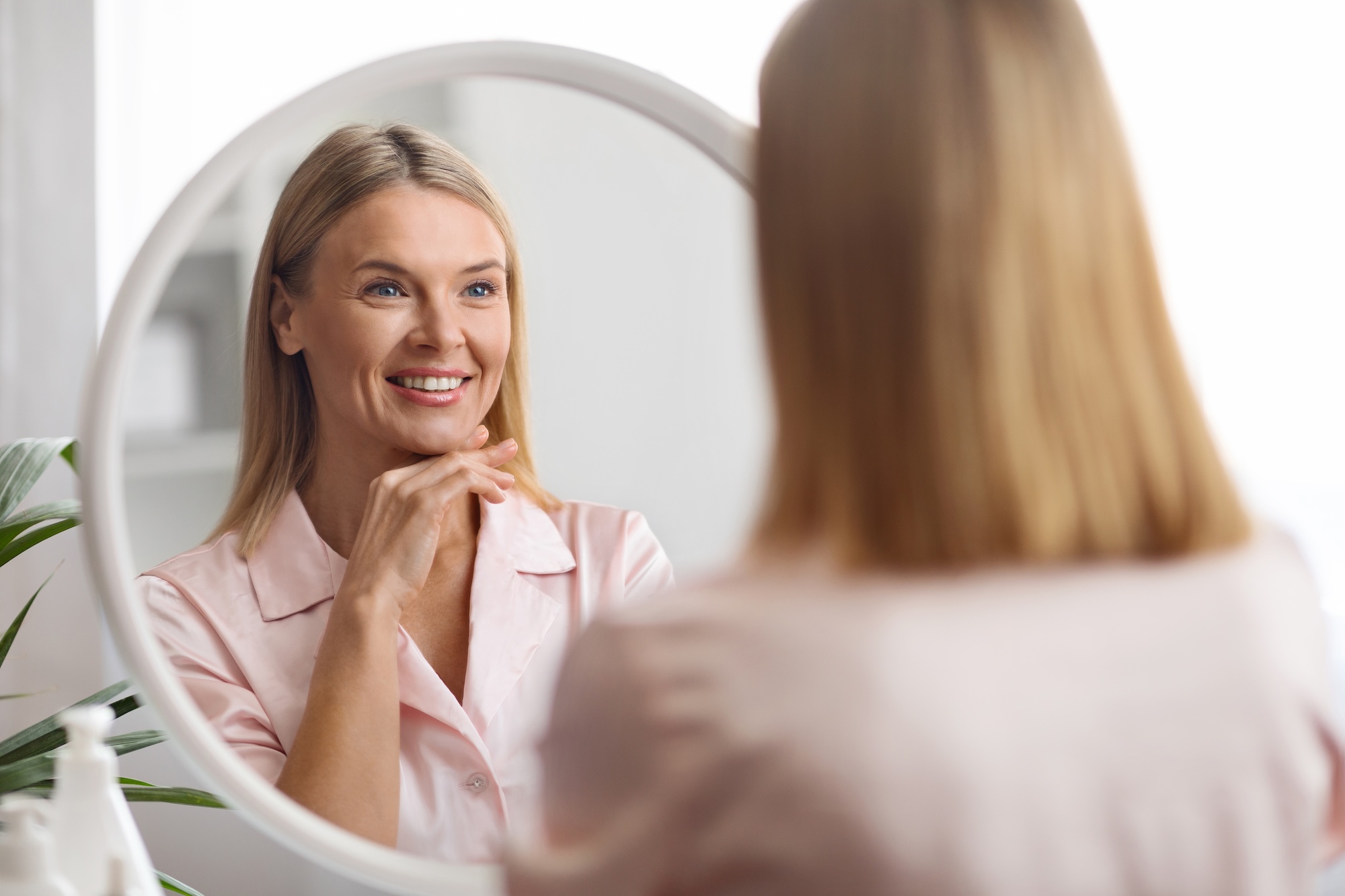 A Blonde woman happily looking in the mirror at home, admiring her facial shape after visiting Vive Aesthetics & Wellness for jawline Botox near Cherry Hill, NJ.