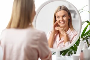 A woman smiles in the mirror after receiving treatment with our best skincare products near Medford.